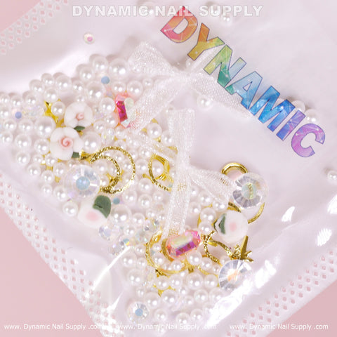 The Luxury White Pearls and Flowers with Leaves Set (White Theme) for Nail Art Design by Dynamic Nail Supply is showcased against a pink background. It includes luxury white pearls, colorful floral beads, and gold rings, all neatly packaged in a clear bag. The brand name "DYNAMIC" gleams in rainbow colors at the top, accompanied by an elegant white lace tied around the center.