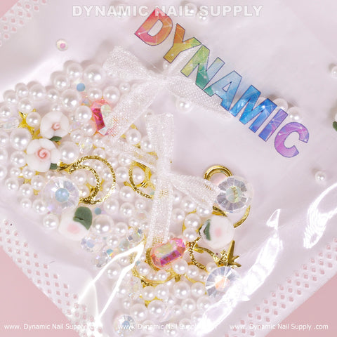 A package from Dynamic Nail Supply showcases their Luxury White Pearls and Flowers with Leaves Set (White Theme) for nail art design, featuring exquisite elements like luxury white pearls, gold crescent moons, pink roses, small bows, iridescent gems, and heart-shaped pieces. These treasures are beautifully displayed beneath the holographic Dynamic Nail Supply brand name on a soft pink backdrop.
