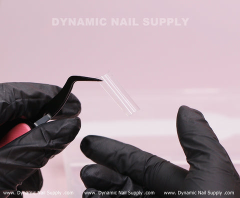 A gloved hand uses tweezers to hold XL Square Nail Tips (Not C-Curve); brand name DYNAMIC NAIL SUPPLY appears behind.