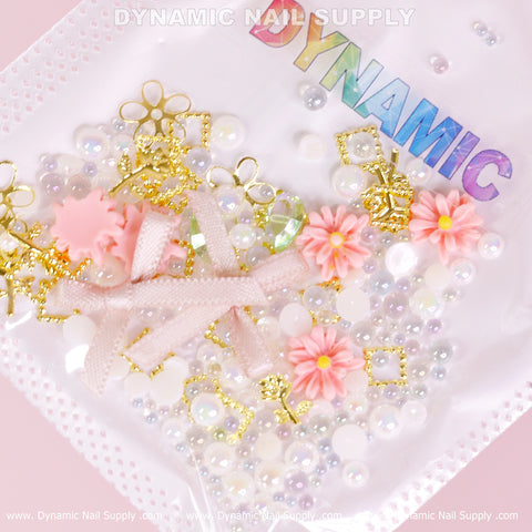 A light pink-themed nail art decoration set from Dynamic Nail Supply features luxury white pearls, colorful flower charms in pink and white varieties, and gold embellishments. The set includes pearl-like beads and iridescent rhinestones for added sparkle. A translucent packet labeled with the brand name showcases the shiny contents of the Luxury White Pearls, Colorful Flowers Set.