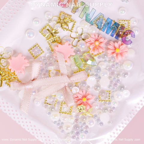 A flat lay showcases an array of nail art decorations from Dynamic Nail Supply, featuring iridescent letters spelling DYNAMIC, complemented by pink bows, colorful flowers, luxury white pearls from the Luxury White Pearls, Colorful Flowers Set (Light Pink Theme) for Nail Art Design, along with gold shapes and silver beads. Arranged on a light pink background, this nail art design exudes playful and creative elegance.