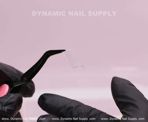 A gloved hand holds tweezers gripping XL Square Nail Tips (Not C-Curve) against a soft pink background.