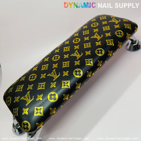 A luxurious manicure service can be enhanced with the (Black Gold LV Pattern) Leather Cushion Armrest with Pillow, featuring yellow monogram-like symbols, perfect for an upscale touch. The armrest by Dynamic Nail Supply is supported by sleek metal legs and is set on a white surface, with the brand's name visible in each corner.