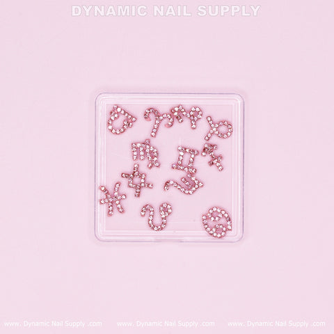 A small clear box contains the Luxury Light Pink Zodiac Symbol Sign Nails Charm Set with rhinestone accents. The charms, shaped like zodiac symbols and letters, include designs such as a star, heart, and dolphin—perfect for enhancing Rhinestone Birthday Nails. The text "Dynamic Nail Supply" is repeated around the border in light gray.