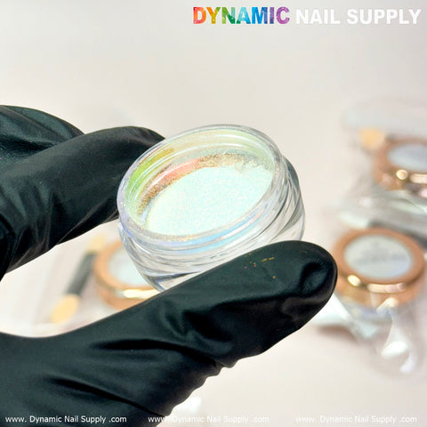 A gloved hand holds a small, clear container filled with Bright-gold/Mermaid Iridescent Chrome #04 - High brightness Fine Pigment Powder. The background features several similar containers softly blurred in the distance. At the top, the brand name Dynamic Nail Supply appears in colorful and grey letters, ideal for your next nail art design.