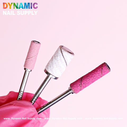 A close-up shows a hand wearing a pink glove and holding three nail drill bits, including the Small 3.5mm Easy-Off Mandrel for Smaller Sanding Band. The drill bits come in various shades and textures against a light pink background. At the top, "Dynamic Nail Supply" is displayed in colorful text, with their website link appearing at the bottom.