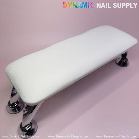 Displayed on a light pink surface, the (White LV Pattern) Full-Grain Leather Armrest Cushion Pillow for Pedicure Manicure by Dynamic Nail Supply features a sleek design elevated on chrome legs. Adding an extra touch of comfort and elegance, this setup highlights Dynamic Nail Supply's name prominently at the top and bottom edges of the image.