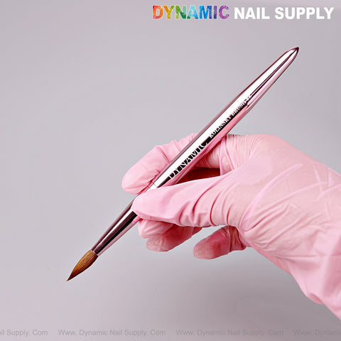 A gloved hand holds the Dynamic Metallic Pink 100% Pure Kolinsky Hair Brush for acrylic nail application.