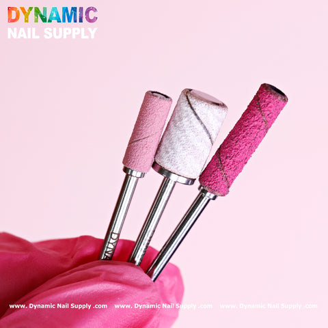 A gloved hand displays three easy-off sanding band mandrels from Dynamic Nail Supply, each in distinct shades and textures of pink, set against a coordinating background. The bold branding of Dynamic Nail Supply is prominently featured in the top-left corner.
