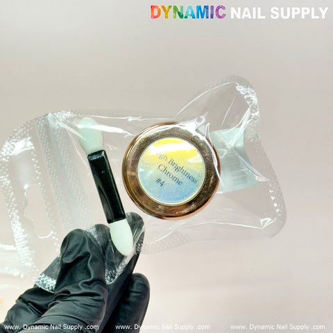 A hand in a black glove holds a sealed package containing a small jar labeled Orange/Gold/Mermaid Iridescent Chrome #06 - High Brightness Fine Pigment Powder and a dual-ended applicator, ideal for nail art. The background features a pale pink surface with the Dynamic Nail Supply brand name at the top.
