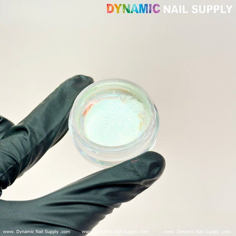 A hand in a black glove holds a small open jar of Light-Mermaid Iridescent Chrome #10 - High brightness Fine Pigment Powder, perfect for creating stunning nail designs. The jar is set against a plain light background, with "Dynamic Nail Supply" displayed in colorful letters at the top right corner.