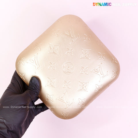 A gloved hand holds the Metallic Gold LV Engrave Elbow Rest Pillow for Nail Tech on a pink background.