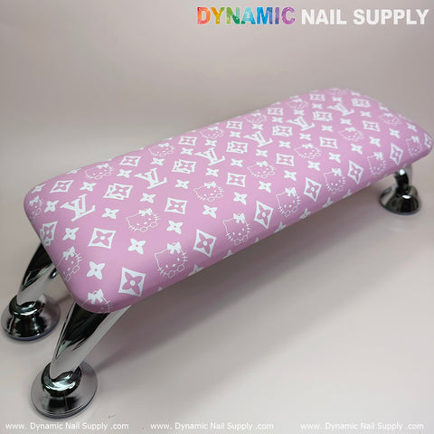 A pink Hell0 Kitty LV nails pillow armrest from Dynamic Nail Supply showcases white patterns, logos, and playful designs resting on shiny silver legs. Paired with a sleek leather cushion for manicure or pedicure sessions, it adds a chic aesthetic against the neutral backdrop. The DYNAMIC NAIL SUPPLY logo is prominently displayed at the top.