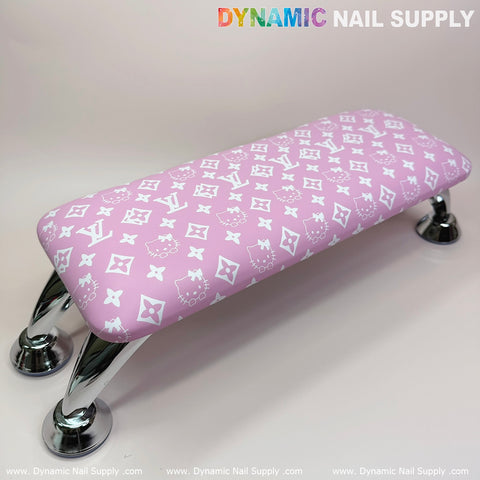 A "Pink Hell0 Kitty LV" nail armrest with a white logo-like pattern provides comfort through its rectangular leather cushion top. Supported by four metallic legs, it is the perfect addition to any manicure and pedicure service setup, with the Dynamic Nail Supply brand prominently featured against a light, neutral background.