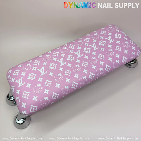 The (Pink Hell0 Kitty LV) Nails Pillow ArmRest from Dynamic Nail Supply features a plush leather cushion adorned with white floral and letter motifs, supported by shiny metal legs, making it ideal for manicure nail services. It rests against a light background with the brand's name elegantly displayed along the top and bottom edges of the image.
