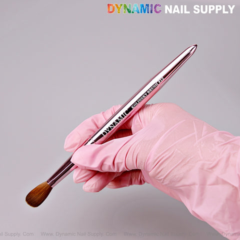A gloved hand holds the Dynamic Metallic Pink 100% Pure Kolinsky Hair Brush for Nails against a light pink background.