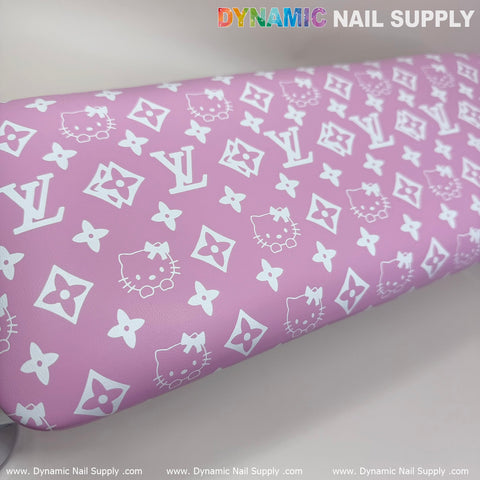 A pink leather cushion adorned with white patterns of interlocking logos, flowers, stars, and cat faces showcases the playful design of the (Pink Hell0 Kitty LV) Nails Pillow ArmRest by Dynamic Nail Supply. Their name is prominently printed at the top, with their website repeated along the bottom. This whimsical and stylish armrest perfectly complements any UV nail light setup for a chic manicure or pedicure service.