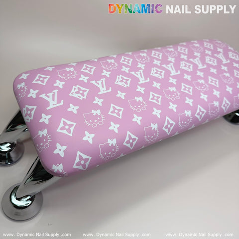 A nail armrest by Dynamic Nail Supply, known as the Pink Hell0 Kitty LV model, features a leather cushion adorned with white floral and logo designs. It is supported by four shiny silver legs and rests against a neutral beige background. The top right corner displays "DYNAMIC NAIL SUPPLY," with their website, www.DynamicNailSupply.com, repeating along the bottom edge.