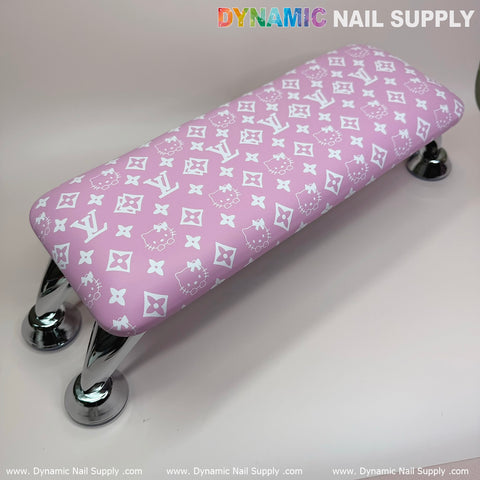 The "Pink Hello Kitty LV" Nails Pillow ArmRest with a Leather Cushion for Manicure Pedicure exudes a luxury brand vibe, featuring a pink design adorned with white logos and cartoon faces. Supported by four shiny chrome legs for stability, it offers enhanced comfort with its leather pillow cushion and is set against a plain background accented with "Dynamic Nail Supply" repeated around the edge.