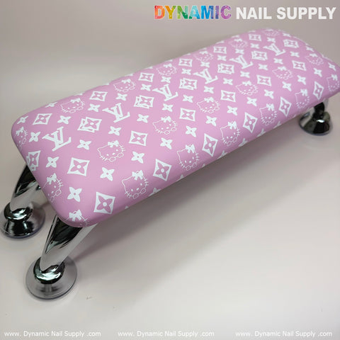 The Pink Hell0 Kitty LV Nails Pillow ArmRest by Dynamic Nail Supply features a stylish design with white logos and emblems on a pink background, supported by shiny curved metallic legs. The top displays a mix of cartoon faces and geometric shapes against a light, neutral backdrop. This leather-cushioned armrest is perfect for your manicure or pedicure service, seamlessly complementing any UV nail light setup.