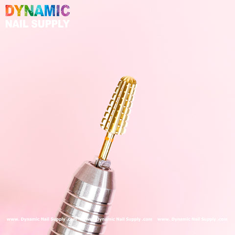 A close-up of a 2XC Volcano Bit, renowned for its superior quality and long-lasting durability when removing acrylic, hard gel, or dipping powder at high speed. The gold tungsten carbide nail drill bit is attached to a metallic holder against a soft pink background. The top left corner features the Dynamic Nail Supply logo in vibrant text, with delicate branding text along the bottom edge.