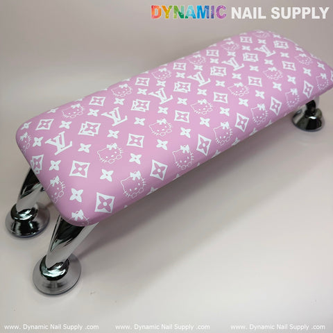 The Pink Hell0 Kitty LV Nails Pillow ArmRest, featuring a leather cushion adorned with a pattern of white Hello Kitty faces and interlocking LV logos, rests on a smooth surface. It is supported by four shiny metal legs for stability during your manicure and pedicure service. The brand name "Dynamic Nail Supply" circles the perimeter of the image.