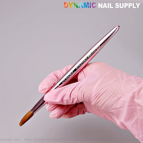 A hand in a pink glove holds the Dynamic Metallic Pink 100% Pure Kolinsky Hair Brush for Nails; logo at top right.