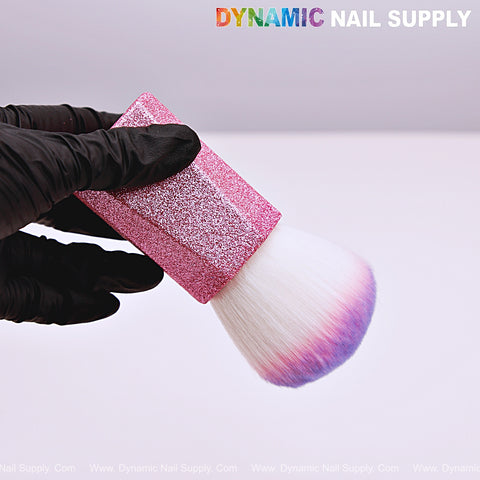 A gloved hand holds the Ombre Pink Glitter Small Dust Brush with purple and blue-tipped bristles against a plain background.