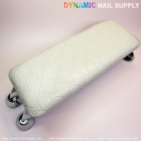 The white, rectangular Armrest with Leather Cushion Pillow from Dynamic Nail Supply features embossed patterns and is supported by four shiny silver legs. It doubles as an armrest for added comfort and showcases intricate, repeated motifs. The neutral and light background highlights the brand name "Dynamic Nail Supply" at the top with a rainbow effect on "Dynamic," making it perfect for enhancing your manicure service experience.