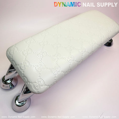 A rectangular, white armrest featuring an embossed leather cushion pillow by Dynamic Nail Supply is supported by four silver chrome feet, making it ideal for any manicure service. The background displays a light pink gradient, with "Dynamic Nail Supply" printed in colorful letters at the top and the website URL repeated along the bottom edge.