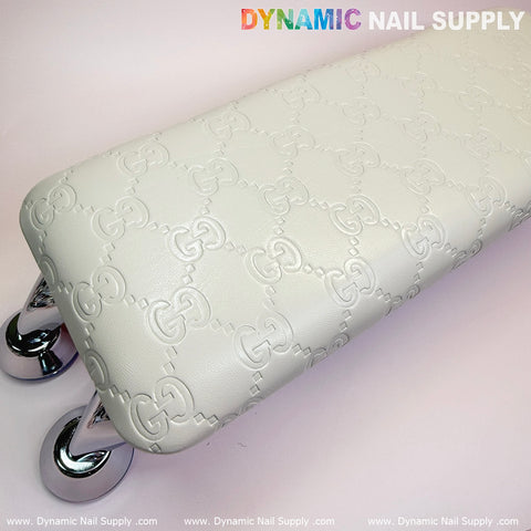 A white armrest with a leather cushion, specifically designed for pedicure and manicure services, features an embossed GC pattern. It is supported by four shiny, silver loop-shaped legs. The background is softly lit with "Dynamic Nail Supply" printed in multicolored text above and "Nail Lamps" in white below.
