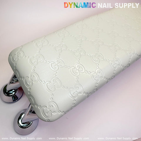 A white armrest embossed with GC logos and featuring a leather cushion pillow for pedicures and manicures is elegantly supported by chrome legs. Ideal for any nail service, it sits gracefully on a light pink surface adorned with the Dynamic Nail Supply brand name and logos around the image's edges.