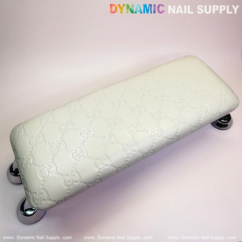 A white GC embossed armrest, featuring a leather cushion pillow ideal for pedicures and manicures, supports a relaxing nail service. It is supported by four shiny, round metallic legs against a simple gradient background. "Dynamic Nail Supply" is prominently displayed at the top in colorful letters near essential nail lamps.