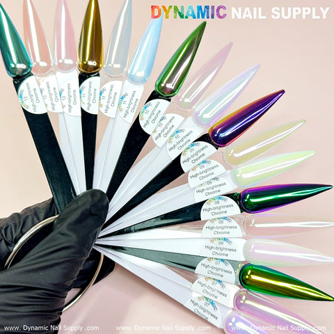 A fan of nail polish swatches featuring different chrome finishes is held in a gloved hand. Each swatch, enriched with the Light-Pearl Transparent Iridescent Chrome #12 - High Brightness Fine Pigment Powder, displays a distinct color reflecting hues such as gold, blue, pink, and green. "Dynamic Nail Supply" is written in the top right corner against a light pink background.