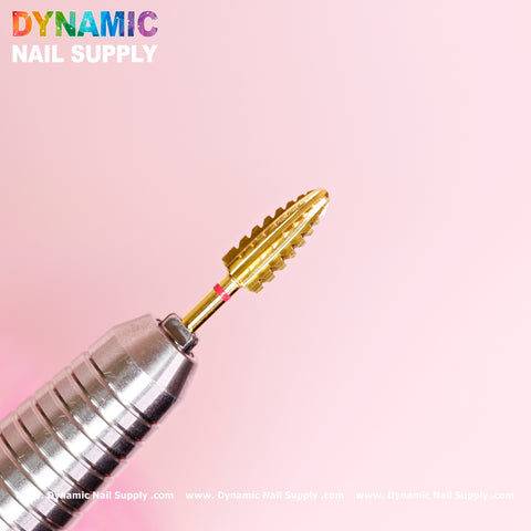 A close-up of the 2XC Typhoon Bit from Dynamic Nail Supply, featuring a gold double extra coarse drill bit with a carved, ribbed design, is attached to a metallic tool against a soft pink background. The brand name "Dynamic Nail Supply," renowned for quality acrylic supplies, is printed in colorful letters at the top left corner. The website information is displayed in white text along the bottom edge.
