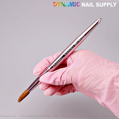 A pink-gloved hand holds the Dynamic Metallic Pink 100% Pure Kolinsky Hair Brush; logo appears on brush and background.