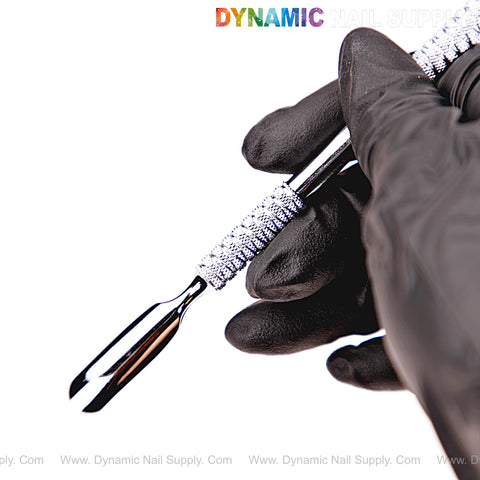 A gloved hand holds the NT1231 – Professional Double-Function Cuticle Pusher (Bevel and Spoon) by Dynamic Nail Supply.