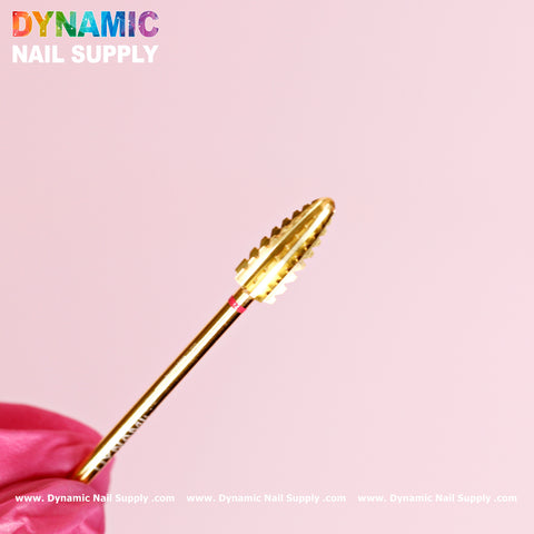 A close-up of the 2XC Typhoon Bit from Dynamic Nail Supply, a double extra coarse drill bit designed for removing acrylic or gel polish, is held by a person wearing a pink glove. Its barrel-shaped, grooved design ensures precise gel polish application. The scene is set against a light pink backdrop, with the brand name Dynamic Nail Supply shimmering in rainbow colors at the top left corner.