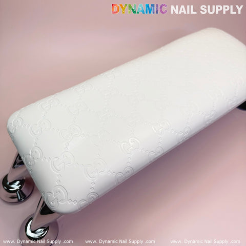 A Super White GC leather armrest with a cushion pillow enhances the surface, featuring a subtle embossed pattern. Its shiny chrome legs add elegance against a light pink background, perfect for any manicure service. The Dynamic Nail Supply brand is prominently displayed at the top, along with their website link below.