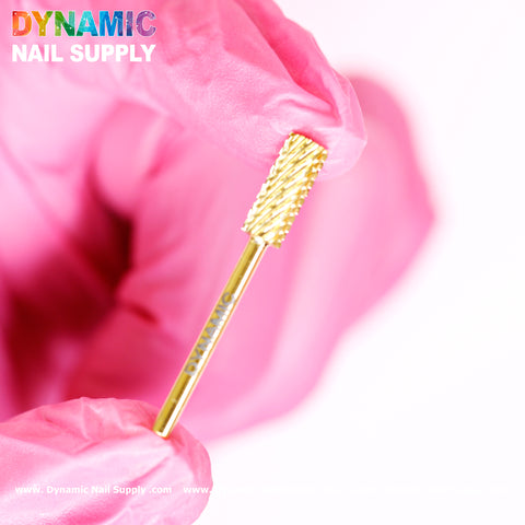 A close-up of a person's hand in pink gloves holding a 2XC Double Extra Coarse Drill Bit - Small Barrel Carbide, ideal for gel or acrylic nail removal. The gold-colored tungsten carbide bit contrasts against the white background, with Dynamic Nail Supply prominently featured at the top left in vibrant text.