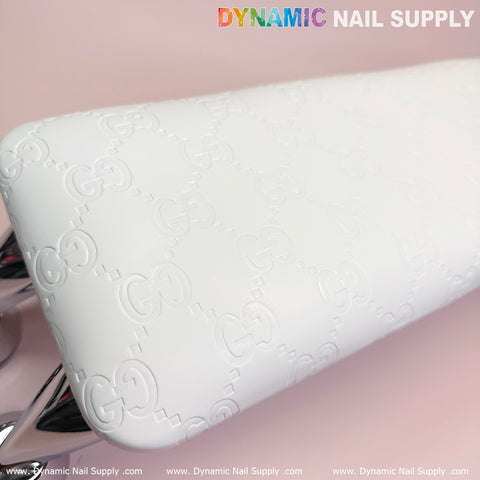 A close-up of the (Super White GC) Leather Armrest with Cushion Pillow for Pedicure Manicure from Dynamic Nail Supply shows its textured design with embossed, repeating letter patterns. Placed on a soft pink surface, the top text features "DYNAMIC NAIL SUPPLY" in a colorful, uppercase font, while the bottom edge displays a web address repeatedly.