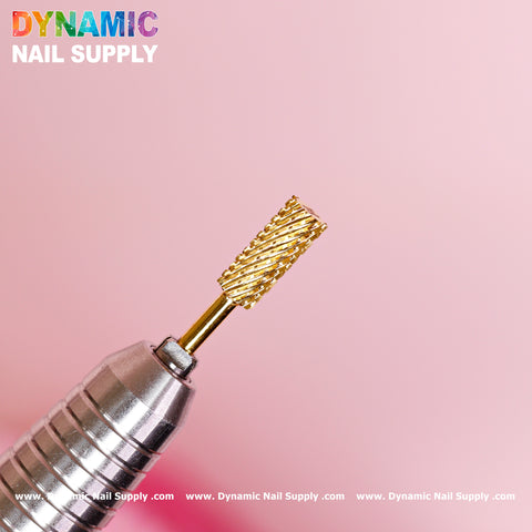 A close-up of a small barrel gold carbide nail drill bit from Dynamic Nail Supply, designed for double extra coarse removal of gel or acrylic nails, is attached to a metallic handle and set against a soft pink background. The drill bit features a textured, ribbed surface, and the brand name "Dynamic Nail Supply" is displayed in a colorful font at the top left corner.