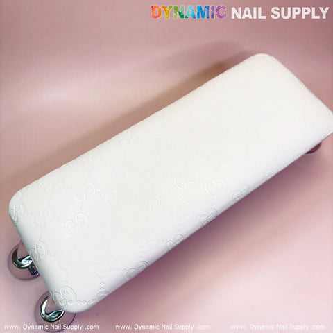 A rectangular armrest in a pristine white shade, slightly embossed with patterns, sits on a soft pink surface, reminiscent of a leather pillow cushion. The armrest features sleek chrome legs. At the top is the brand name "Dynamic Nail Supply" printed in colorful and gray letters, accompanied by the website repeated along the bottom edge.