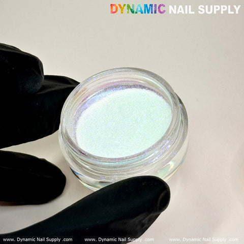 A close-up of a small jar containing Light-Pearl Transparent Iridescent Chrome #12 - High brightness Fine Pigment Powder with a pearlescent sheen. The jar, ideal for creative nail art, is held by a hand wearing black gloves. Text at the top reads Dynamic Nail Supply. The background is plain and light-colored.