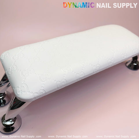 A (Super White GC) Leather Armrest with Cushion Pillow for Pedicure Manicure from Dynamic Nail Supply, featuring a subtle embossed pattern, is supported by chrome legs beside a UV nail light against a soft pink background. In the top right corner, the text "DYNAMIC NAIL SUPPLY" is displayed in multicolored and gray. The bottom edge showcases the website URL repeatedly.