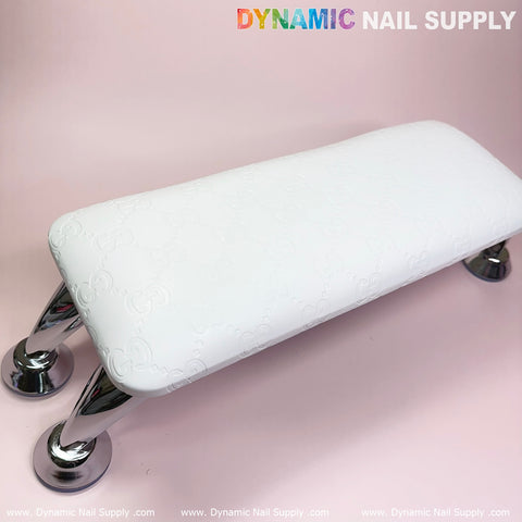 A (Super White GC) Leather Armrest with Cushion Pillow for Pedicure Manicure by Dynamic Nail Supply, resembling a sleek leather pillow cushion, stands on four shiny metal legs against a soft pink background. The surface features a subtle embossed pattern. The brand name Dynamic Nail Supply is displayed in the upper right corner in colorful block letters.