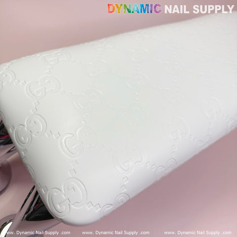 Close-up of the (Super White GC) Leather Armrest with Cushion Pillow for Pedicure Manicure, featuring intricate embossed patterns resembling interlocking letters. The top edge showcases the Dynamic Nail Supply logo in vibrant colors. A UV nail light emits a gentle glow on the pink background, partially visible on the left side and around the armrest.