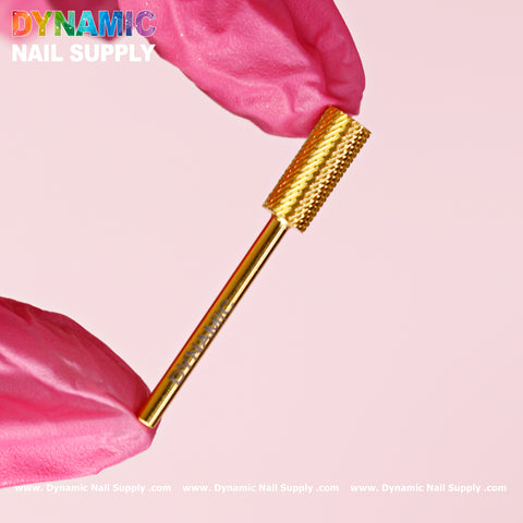 A close-up of Dynamic Nail Supply's Medium Grit (M) Small Barrel bit, made from gold tungsten carbide, is carefully held between fingers adorned with pink gloves. The bit showcases a textured, cylindrical surface specifically crafted for smoothing or shaping nails. A soft pink background accentuates the colorful Dynamic Nail Supply logo positioned at the top left corner.