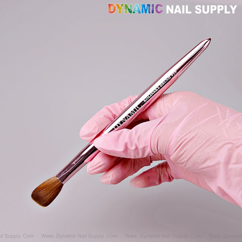 A pink-gloved hand holds a Dynamic Metallic Pink Kolinsky Hair Brush for nails against a light background.