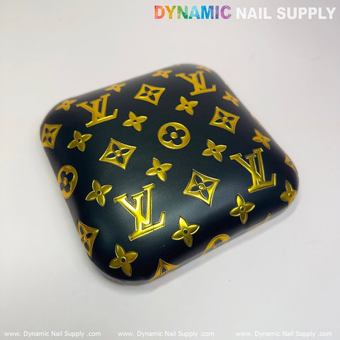 A square elbow cushion pad made from PU leather showcases a dark background adorned with yellow floral motifs and stylized initials, echoing luxury brand designs. It sits elegantly on a light surface, topped with the brand name "Dynamic Nail Supply" in vibrant multicolored text—ideal for any manicure nail desk setting.
